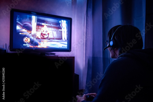 Young male gamer with glasses and headset playing video game at home in the dark room using game console controller watching at LED TV. Gaming and entertainment concepts