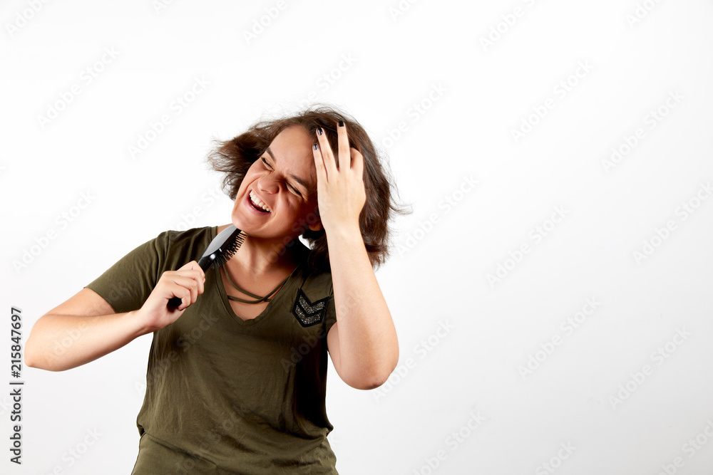 Young brunette woman with tangled unruly hair in casual khaki shirt holding comb and pressing palm to head in pain with closed eyes and open mouth standing over white studio background Young brunette woman with tangled unruly hair in casual khaki shirt holding comb and pressing palm to head in pain with closed eyes and open mouth standing over white studio background