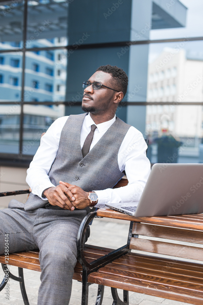 african businessman in suit and glasses is sitting at a table with a Laptop, a cup of coffee against a background of glass buildings african businessman in suit and glasses is sitting at a table with a Laptop, a cup of coffee against a background of glass buildings