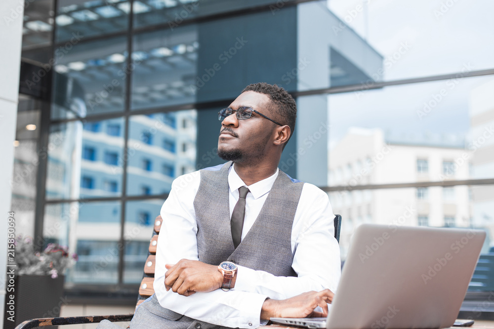 african businessman in suit and glasses is sitting at a table with a Laptop, a cup of coffee against a background of glass buildings african businessman in suit and glasses is sitting at a table with a Laptop, a cup of coffee against a background of glass buildings