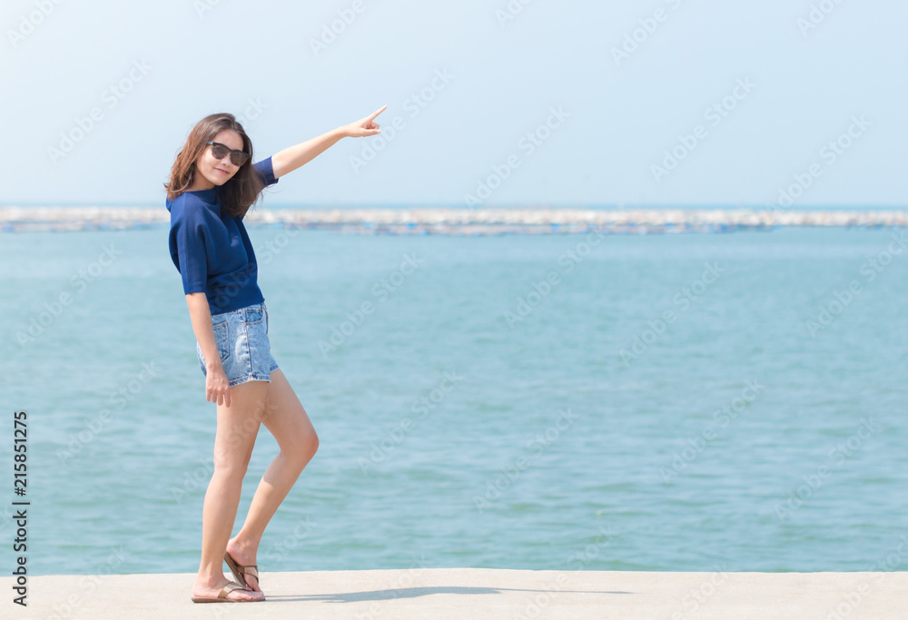 Full length of Asian woman in sunglasses, blue casual and jean shorts standing and pointing her finger to the sky. vacation at sea. Full length of Asian woman in sunglasses, blue casual and jean shorts standing and pointing her finger to the sky. vacation at sea.