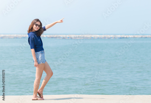 Full length of Asian woman in sunglasses, blue casual and jean shorts standing and pointing her finger to the sky. vacation at sea.