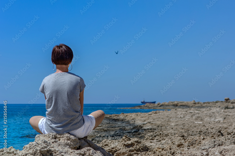 Dziewczyna siedząca na skale nad morzem, patrząca na wrak statku MV Demetrios II, Paphos, Cypr - obrazy, fototapety, plakaty 