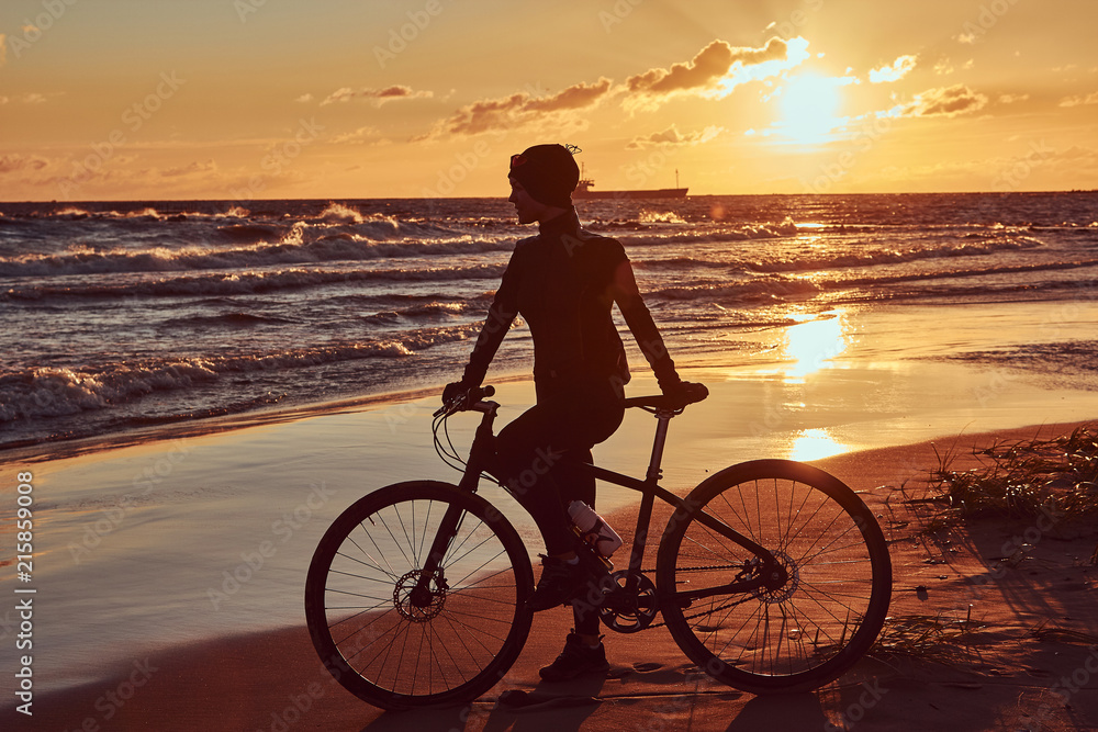 Fototapeta premium Female cyclist standing with her bicycle and enjoying the sunset on the sea coast.