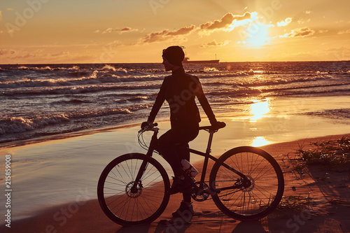 Female cyclist standing with her bicycle and enjoying the sunset on the sea coast.