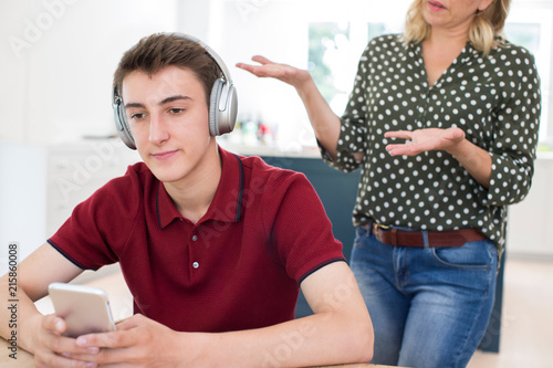 Fotografi Teenage Boy Wearing Headphones And Using Mobile Phone Being Nagged By Mother At
