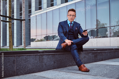Portrait of a confident stylish businessman dressed in an elegant suit holds a smartphone and looking at camera while sitting outdoors against a skyscraper background.