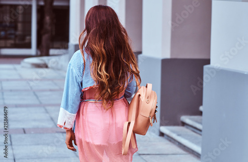 Back view of a woman with long brown hair dressed in trendy clothes holds a rucksack while standing on the street.