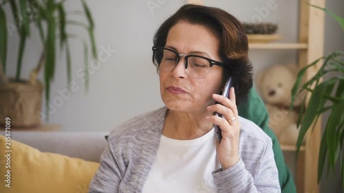 Mature woman in glasses with brown hair sitting on the sofa with yellow pillow and talking on phone with. Indoors. Portrait.