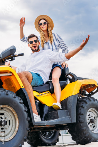 happy young couple sitting on ATV in front of cloudy sky and looking at camera