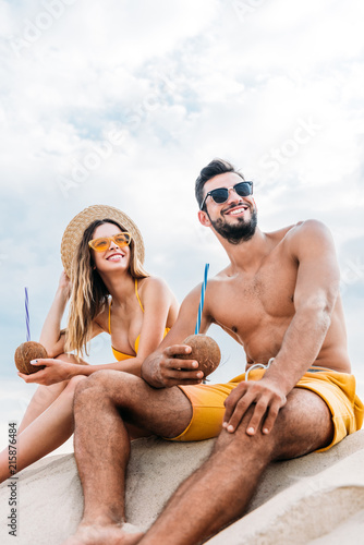 bottom view of young couple with coconut cocktails sitting on sand in front of cloudy sky