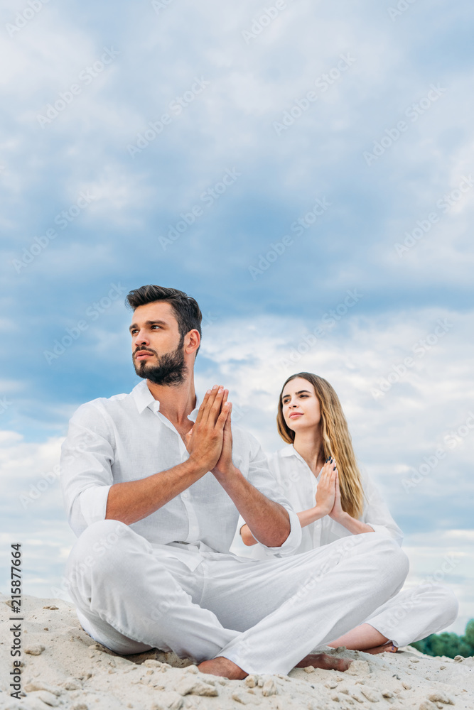 beautiful young couple practicing yoga while sitting on sandy dune in lotus pose (padmasana) and looking away
