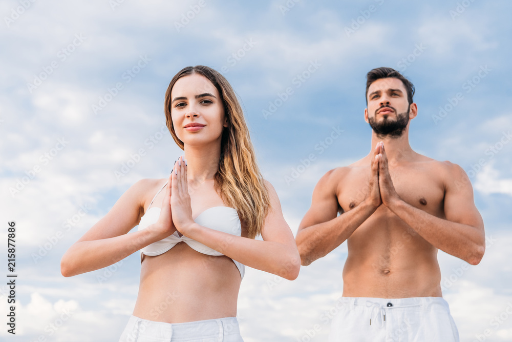 Young couple making namaste mudra and practicing yoga in front of cloudy sky