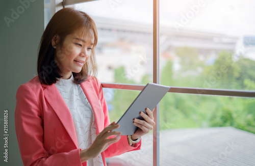 Free Download Attractive businesswoman wear pink suit using a digital tablet while standing in front of windows in an office building and sun light. - GFXLoad
