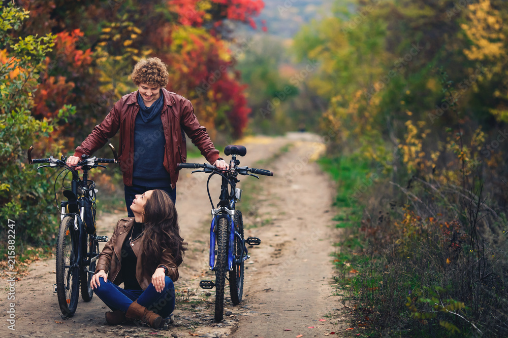 Naklejka premium curly-haired mustachioed guy and an ore-haired girl in autumn stand with bicycles on road and show cheerful emotions.