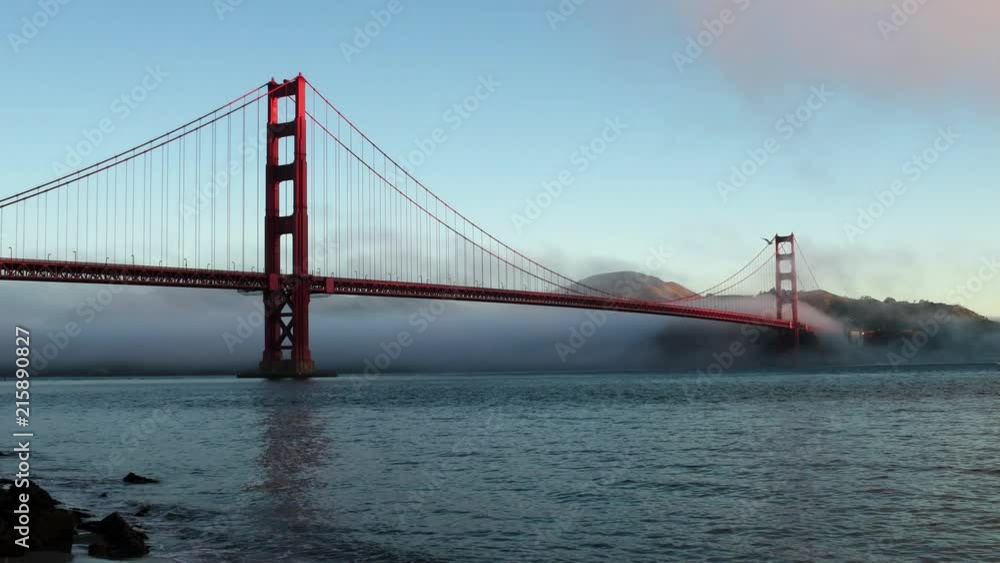 Early morning view of the Golden Gate Bridge, looking west from the Warming Hut. Low fog behind the bridge, light blue sky. Morning sun tinting the top of the towers. Early morning view of the Golden Gate Bridge, looking west from the Warming Hut. Low fog behind the bridge, light blue sky. Morning sun tinting the top of the towers.