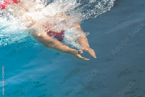 Swimmer's hands just under the surface of the water, trailing bubbles.  Clear, deep aqua blue water.  Red swim cap and suit partially visible below surface waves and foam.