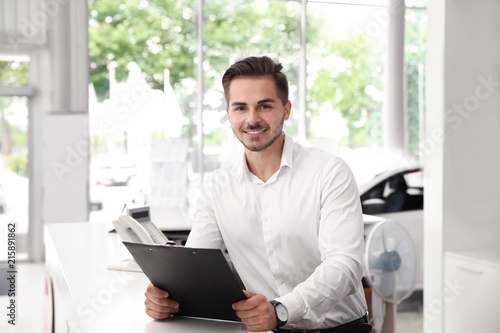 Young salesman with clipboard at desk in car salon