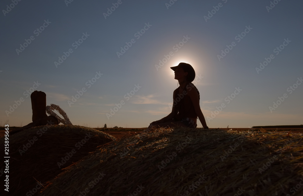 A young woman dressed as a cowgirl holds a straw heart in her hand. She takes time out and enjoys the summer sun  setting as she leisures on a hay bale.