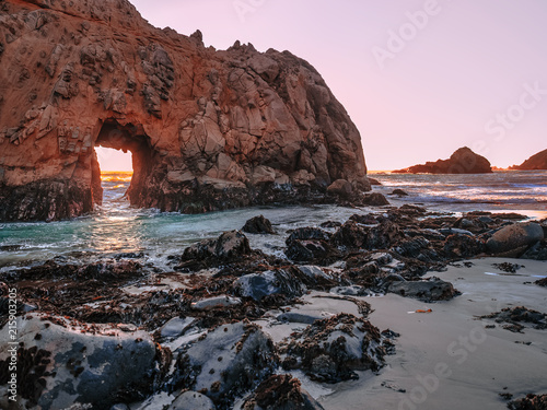 Panoramic view of the Keyhole Arch cliff at Pfeiffer Big Sur State Park beach in California. Black rocks in the foreground. Light of the setting sun creates an amazing red glow on the horizon. photo