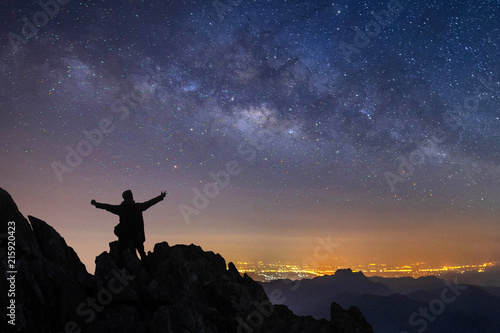 silhouette of a standing man on top of a cliff with arms raised at night landscape mountain and milky way galaxy background , Thailand , long exposure ,low light