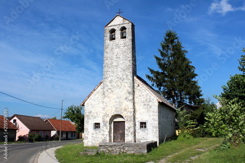 Stone church with tall stone bell tower next to paved road and small family houses surrounded with uncut grass with cloudy blue sky in background