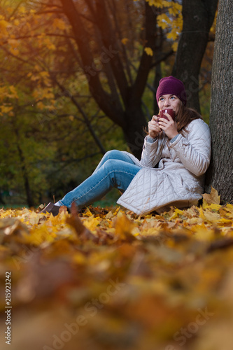 Autumn concept - Young beautiful woman wearing coat and knitted hat sitting with coffee mug on fall nature background