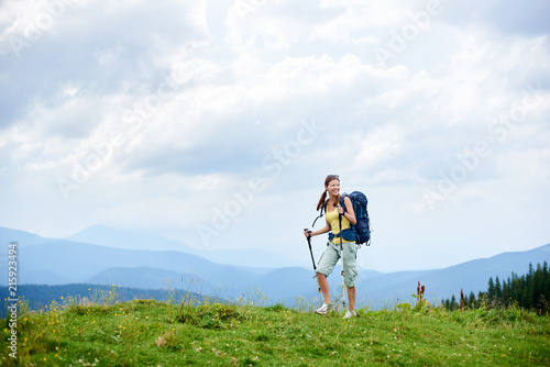 Sporty smiling woman backpacker hiking in Carpathian mountain trail, walking on grassy hill, wearing backpack, using trekking sticks, enjoying summer cloudy day. Outdoor activity, lifestyle concept