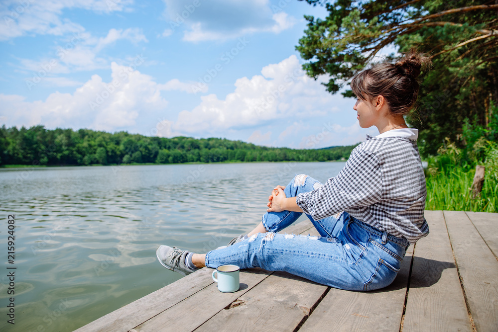 young adult woman sitting on wooden dock drinking coffee and looking at lake