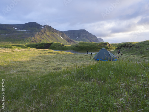 small blue tent standing alone on green grass on mossed creek banks in Hornstrandir Iceland, snow patched hills and cliffs, cloudy sky background, golden hour light, copy space