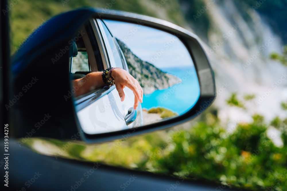Tanned female hand in the car side view mirror. Blue mediterranean sea and white rocks landscape in background
