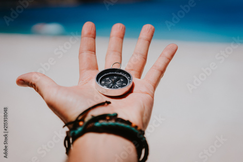 Open palm with stretched fingers holding black metal compass against white sandy beach. Find your way or goal concept. Point of view pov photo