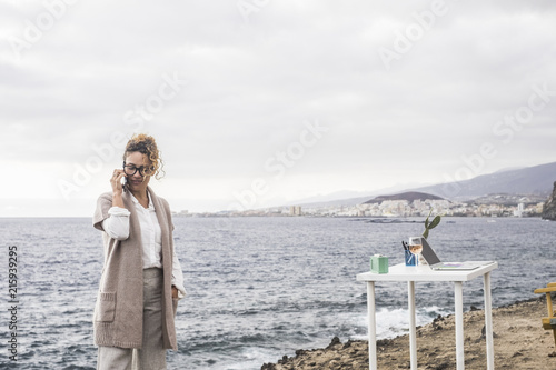 beautiful lady at work with mobile phone and laptop on the desktop in alternative office outdoor in front of the ocean. calling for business. elegant dress and serious hair. wine on the table photo