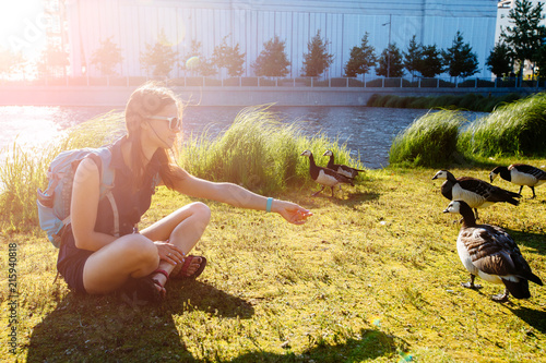 Young tourist woman in sunglasses sitting on the grass and feeding birds Barnacle Goose near the lake. Norway trip in Oslo city concept. Sun glare effect. photo