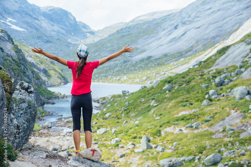 Adventure, travel, tourism, hike and people concept - cheering woman in pink t-shirt open arms over mountains nature background. Female admiring views, relaxing during climbing on Kjeragbolten, Norway