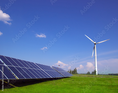 wind generator of electricity from three blades and solar panels of a battery of photocells against a background of clouds and a blue sky green grass