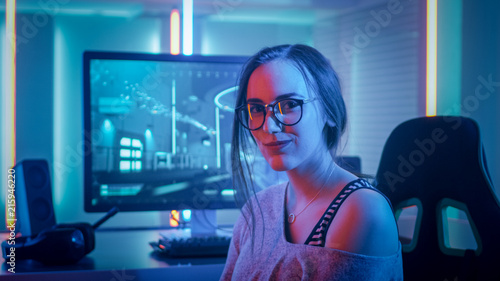 Portrait of the Beautiful Young Pro Gamer Girl Sitting at Her Personal Computer and Looks into Camera. Attractive Geek Girl Player Wearing Glasses in the Room Lit by Neon Lights. photo