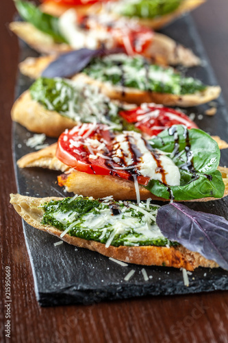 Italian bruschettes with mozzarella cheese, tomatoes, pesto sauce and parmesan cheese, on a black stone, on a wooden table. Copy space, selective focus