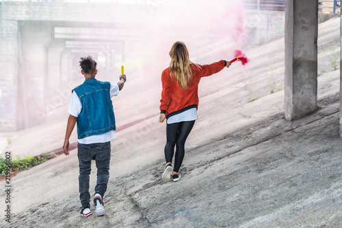 rear view of stylish couple holding colorful smoke bombs at city street