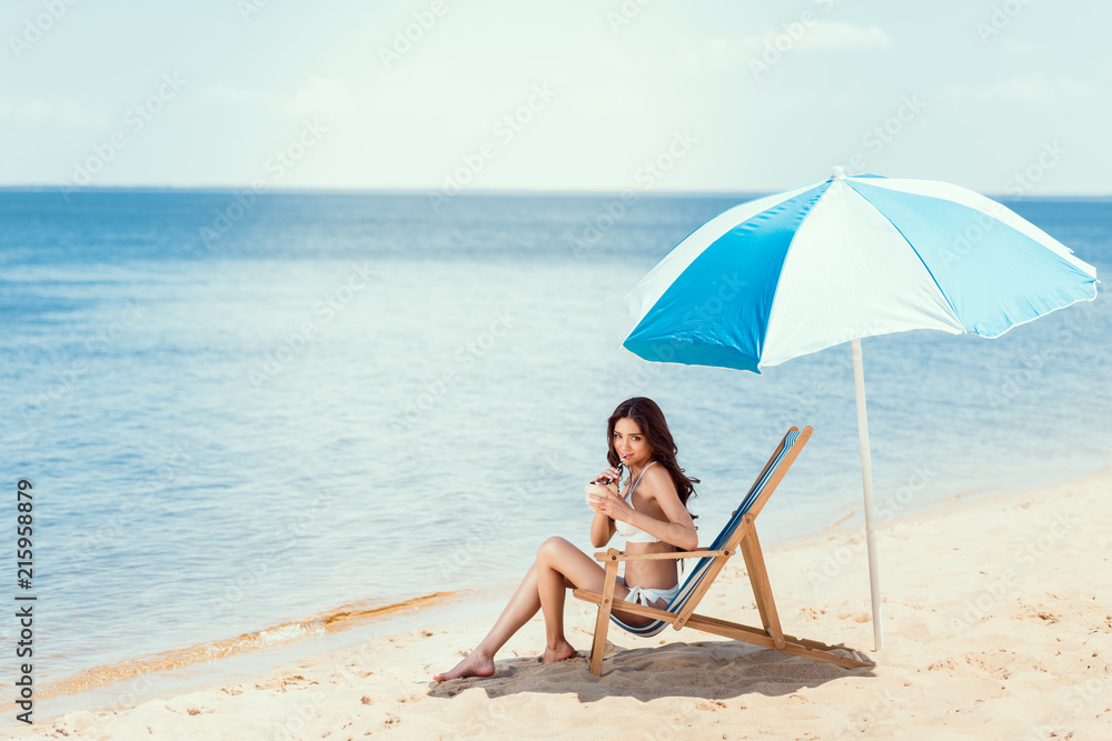 young woman in bikini drinking coconut cocktail in beach chaise longue under sun umbrella near sea young woman in bikini drinking coconut cocktail in beach chaise longue under sun umbrella near sea