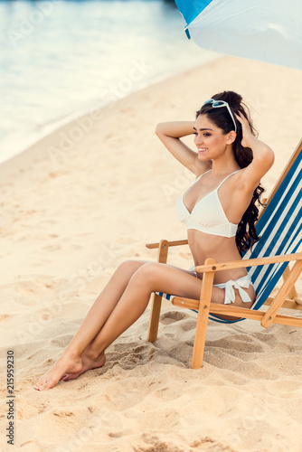 happy brunette girl sitting on beach chair under umbrella near the sea