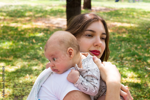 Young mother caring her cute baby on the shoulder outside in the park during nice sunny day, Infant head resting on the shoulder in burping position, mom holding newborn