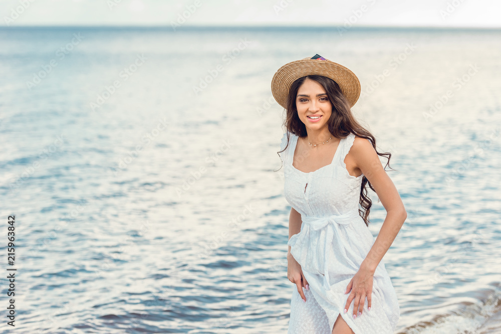 smiling woman in straw hat and white dress walking near the sea