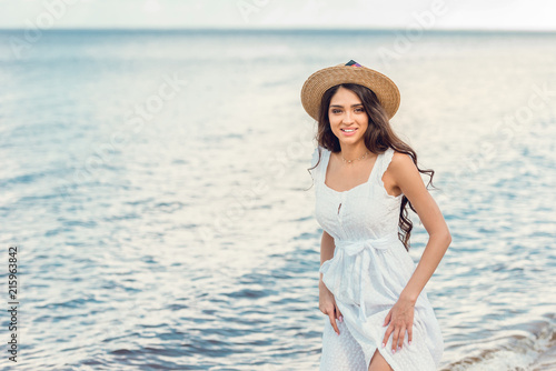 smiling woman in straw hat and white dress walking near the sea