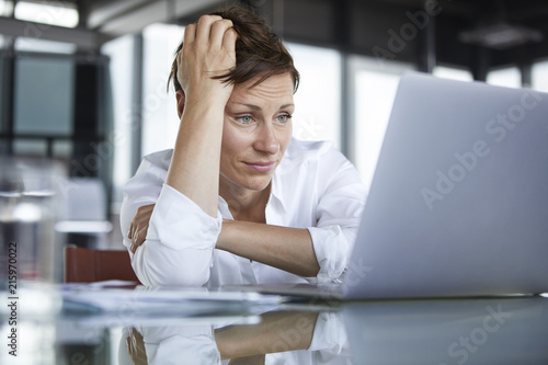 Frustrated businesswoman sitting at glass table in office looking at laptop