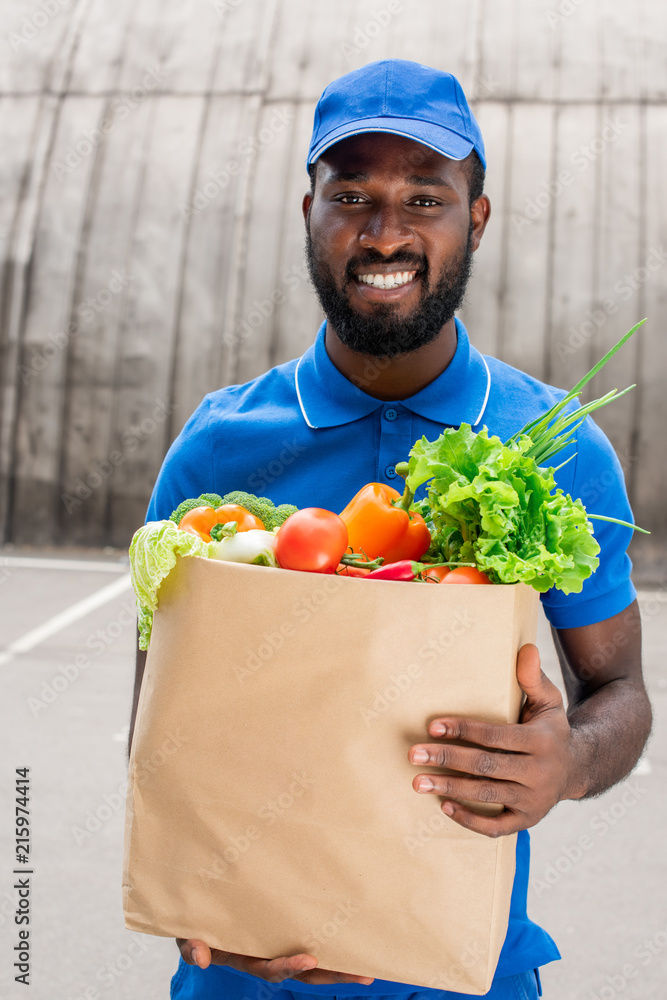 smiling african american delivery man holding paper bag with vegetables smiling african american delivery man holding paper bag with vegetables