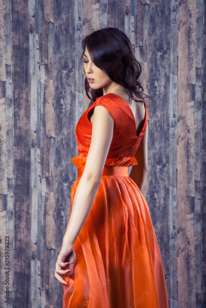 Fototapeta premium Young brunette woman in stylish orange silk dress posing on wooden background