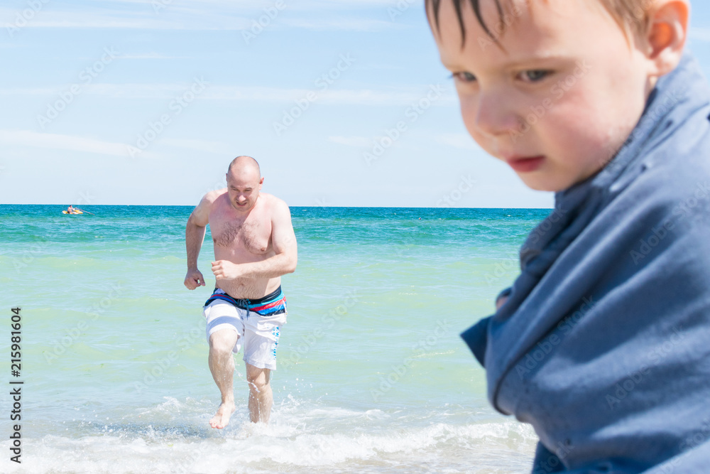 Dad runs out of the water while his son is on the beach.