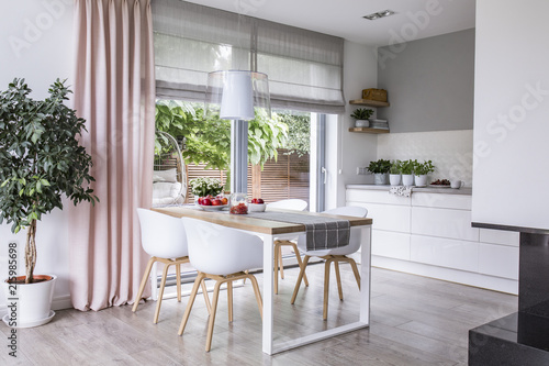 Gray roman shades and a pink curtain on big, glass windows in a modern kitchen and dining room interior with a wooden table and white chairs photo