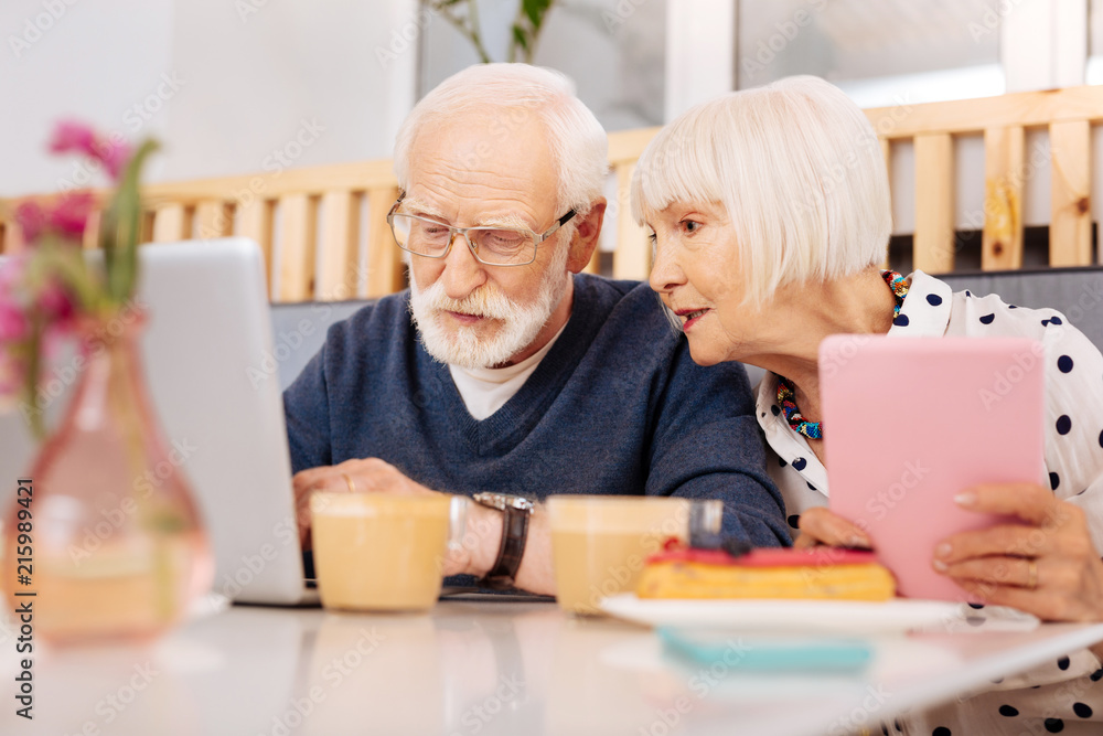Online service. Low angle of pensive senior man using laptop and senior woman grasping tablet